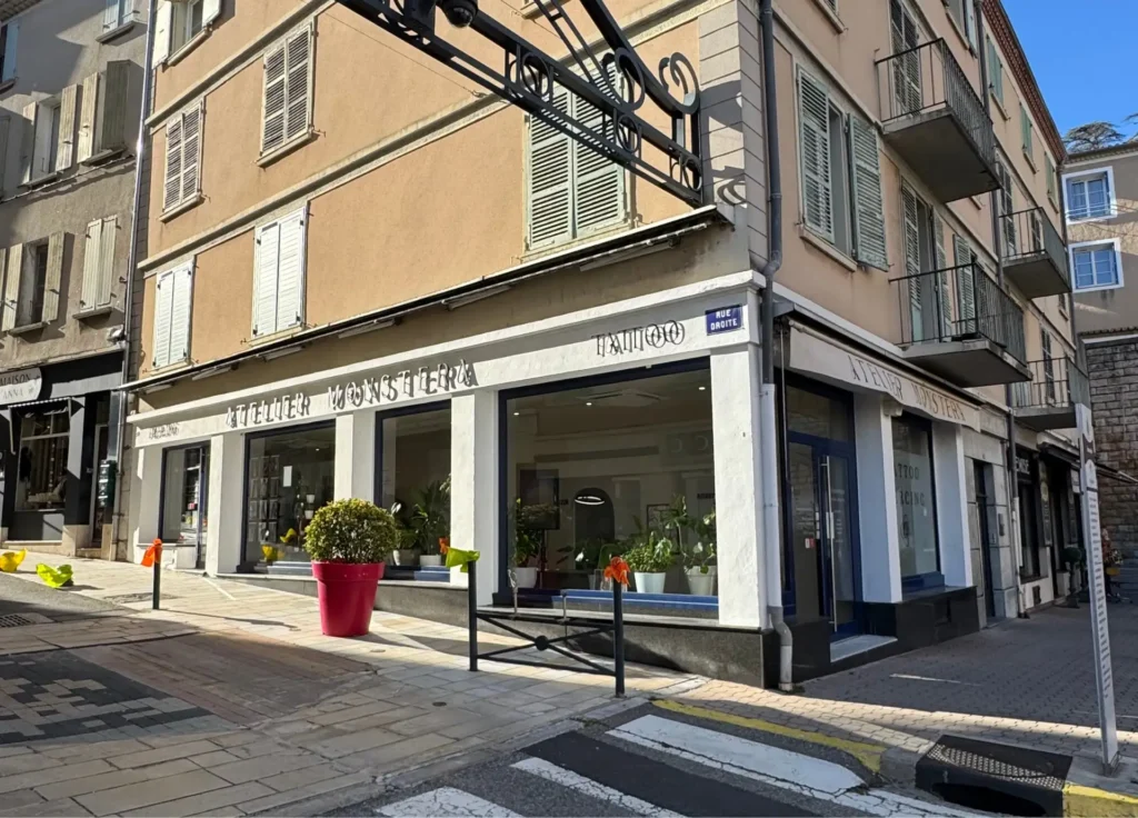 Façade du magasin "Atelier Monstera Tattoo" avec une enseigne lumineuse, des plantes vertes en pots et des fenêtres donnant sur l'intérieur.