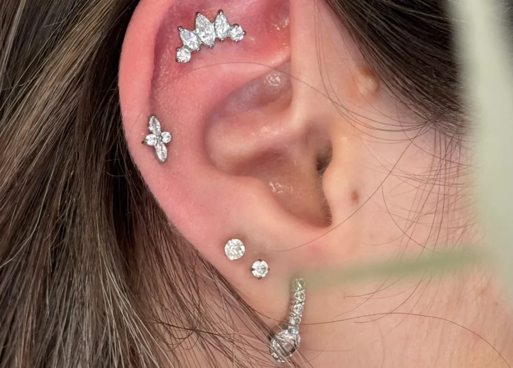 Oreille droite ornée de plusieurs piercings élégants : un bijou en forme de croix, un cluster de cinq pierres en arc-de-cercle, deux clous sertis d'une pierre, et une bague d'oreille sertie de diamants.