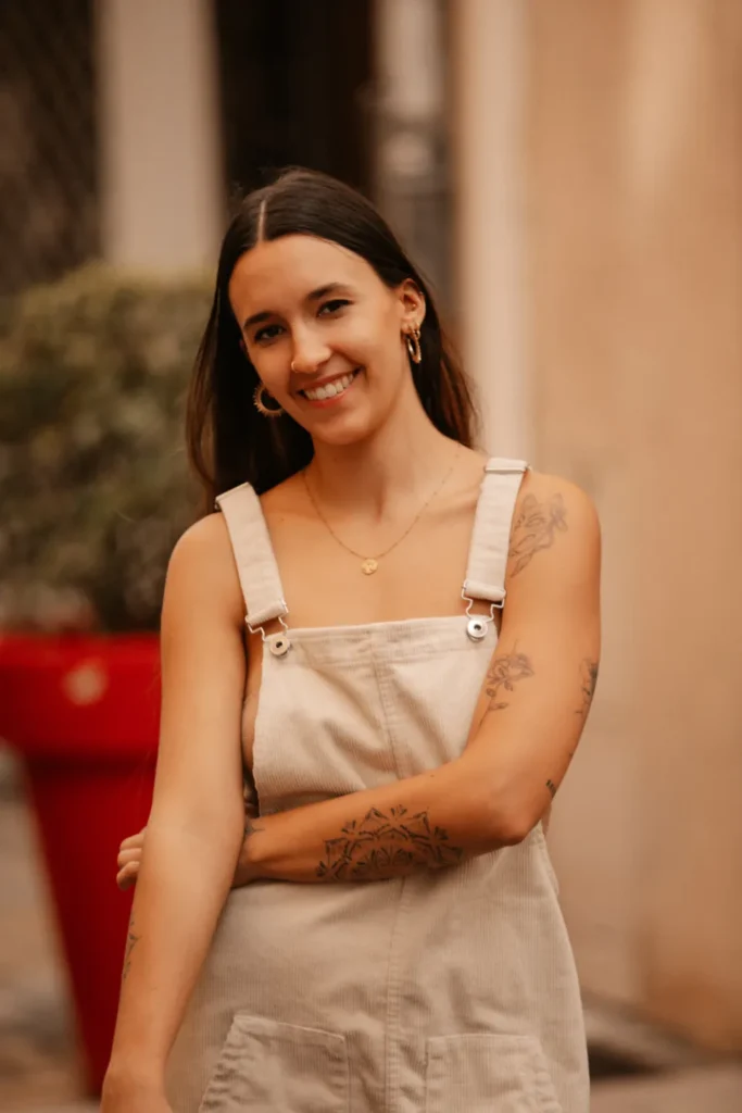 Jeune femme souriante portant une salopette en velours côtelé beige, avec des tatouages sur les bras et des bijoux discrets.