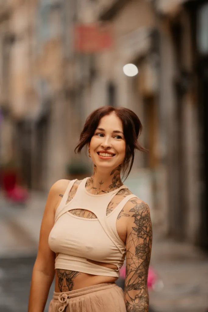 Jeune femme souriante avec des tatouages sur le cou, les bras et le torse, portant un haut beige découpé et un pantalon ample, dans une rue pavée.
