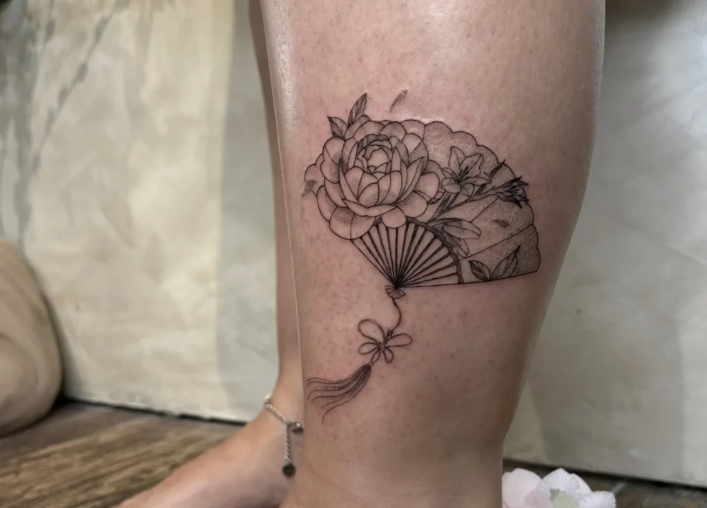 Tatouage réaliste d'un éventail orné de pivoines et de lys, avec un pompon décoratif suspendu.