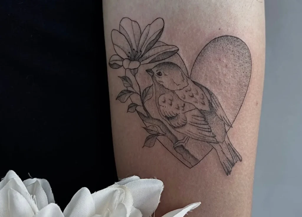 Tatouage d'un oiseau sur une branche devant une fleur, le tout sur fond de cœur texturé.