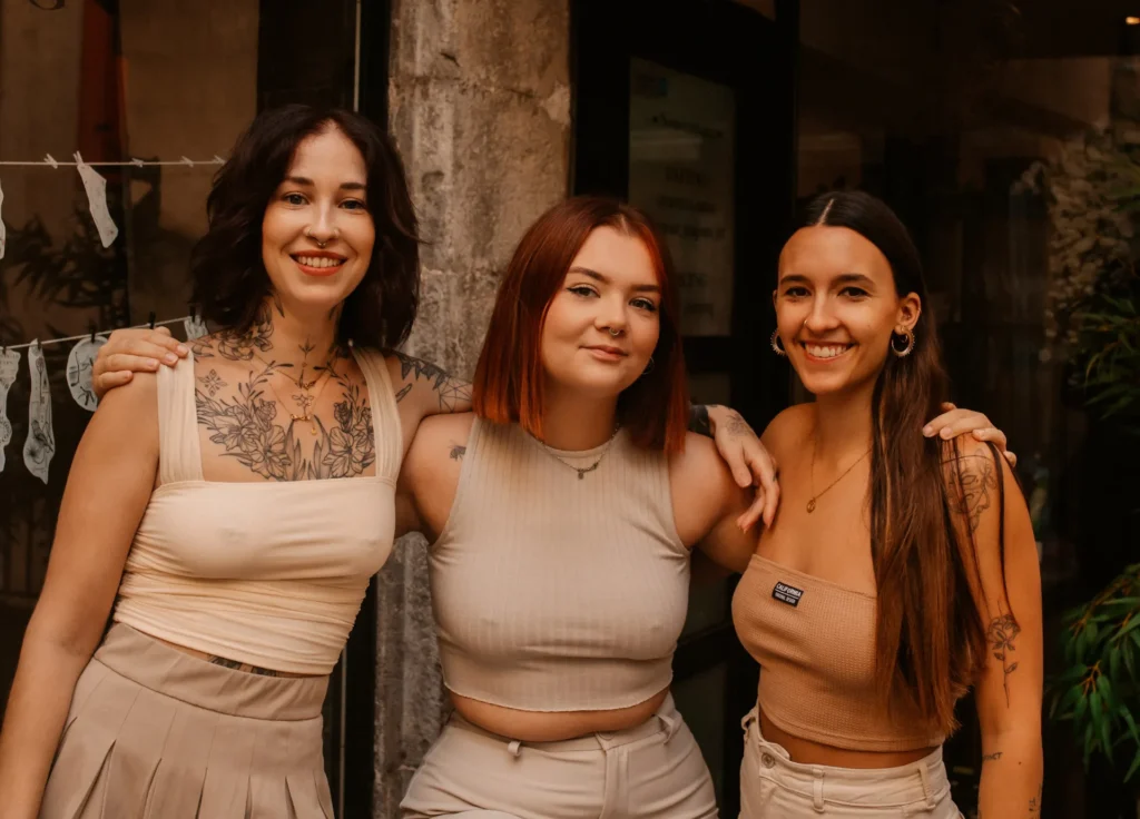 Trois jeunes femmes souriantes se tiennent épaule contre épaule. Elles portent des hauts courts et des pantalons ou jupes de couleur neutre. La femme de gauche a des tatouages sur le torse et le cou. La femme du milieu a les cheveux roux courts. La femme de droite a de longs cheveux bruns et des boucles d'oreilles.