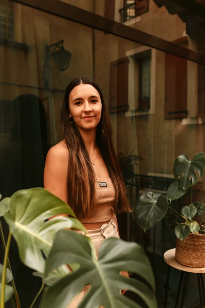 Une jeune femme souriante aux longs cheveux bruns pose devant une fenêtre reflétant un bâtiment avec des volets et une lanterne.