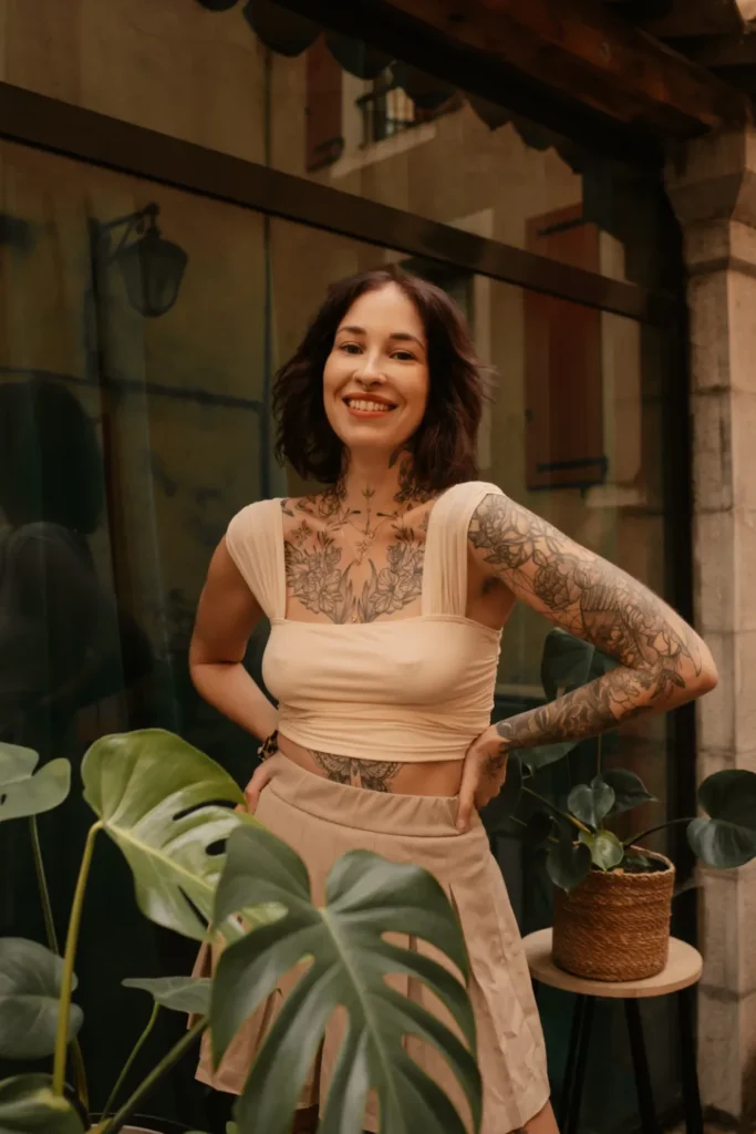 Jeune femme souriante avec de nombreux tatouages sur le corps, portant un haut beige et une jupe, entourée de plantes vertes.