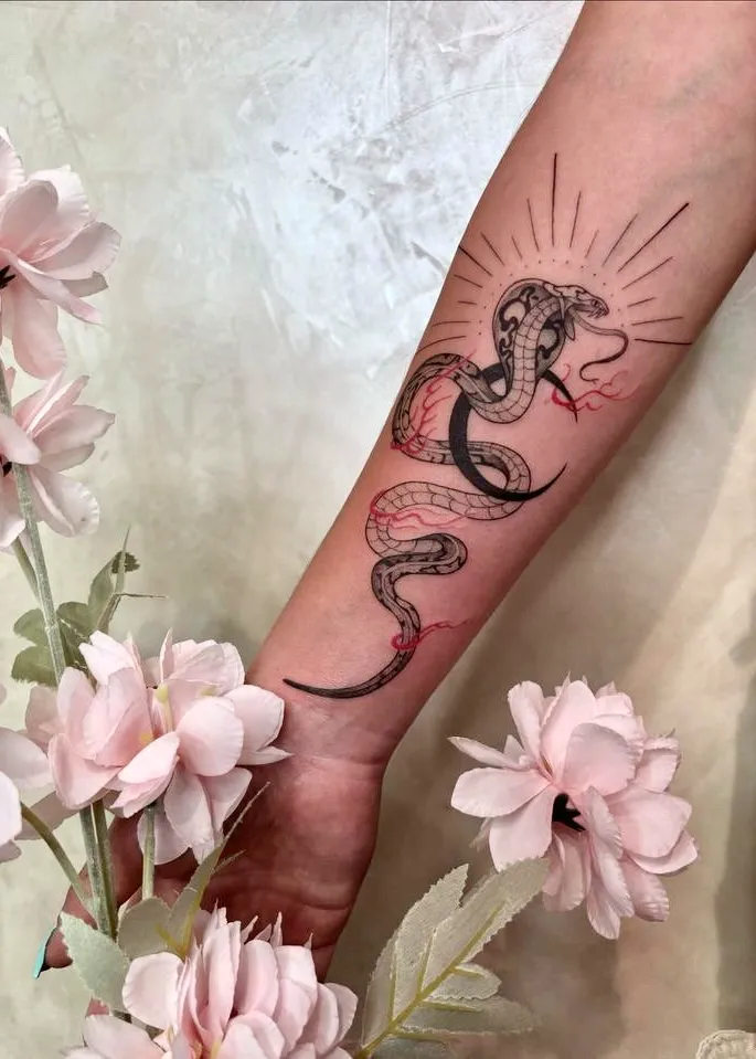 Tatouage de cobra enroulé sur l'avant-bras, orné de détails rouges et d'un style illustratif, avec des fleurs roses en premier plan.