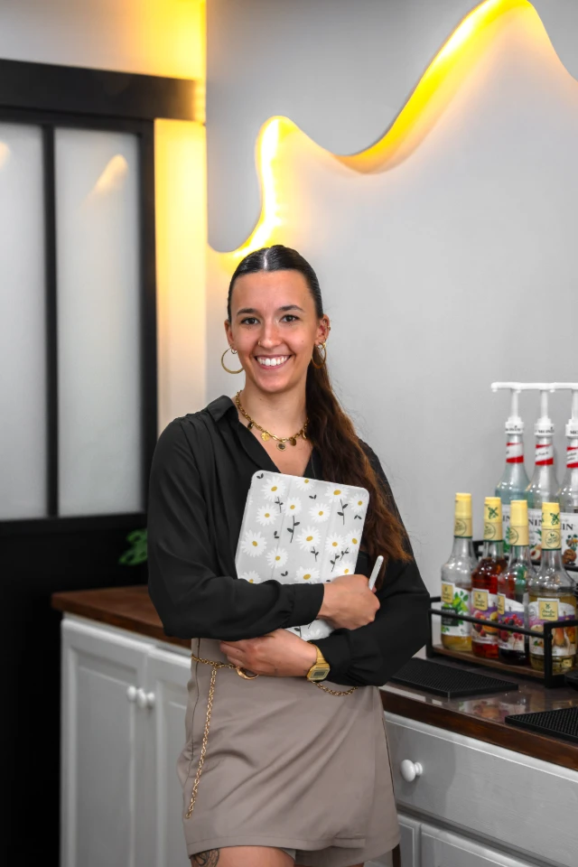 Jeune femme souriante, habillée en tenue de barista, tenant une tablette avec un motif de marguerites.