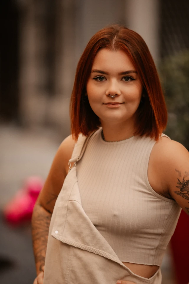 Portrait d'une jeune femme aux cheveux roux et courts, portant un bijou au nez et des tatouages visibles sur ses bras.