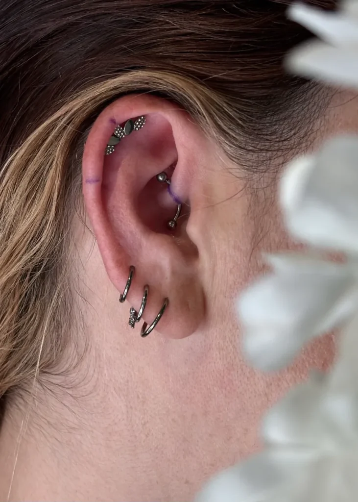 Oreille gauche ornée de plusieurs piercings, dont une courbe de petits ronds sur le cartilage et trois anneaux foncés au lobe, un avec une petite pierre scintillante.