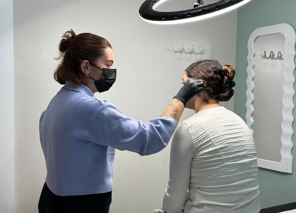 Une perceuse porte des gants noirs et un masque noir, ajustant l'oreille d'une cliente assise devant elle.