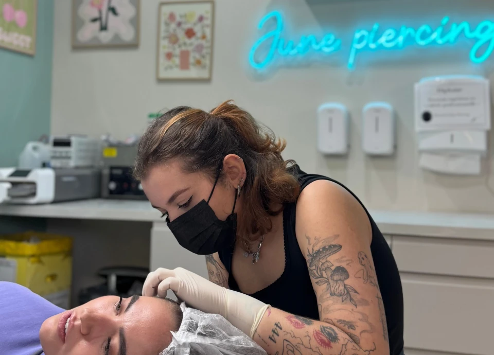 Une pierceuse portant un masque et des gants examine le nez d'une cliente allongée, avec un néon 'June Piercing' au-dessus.