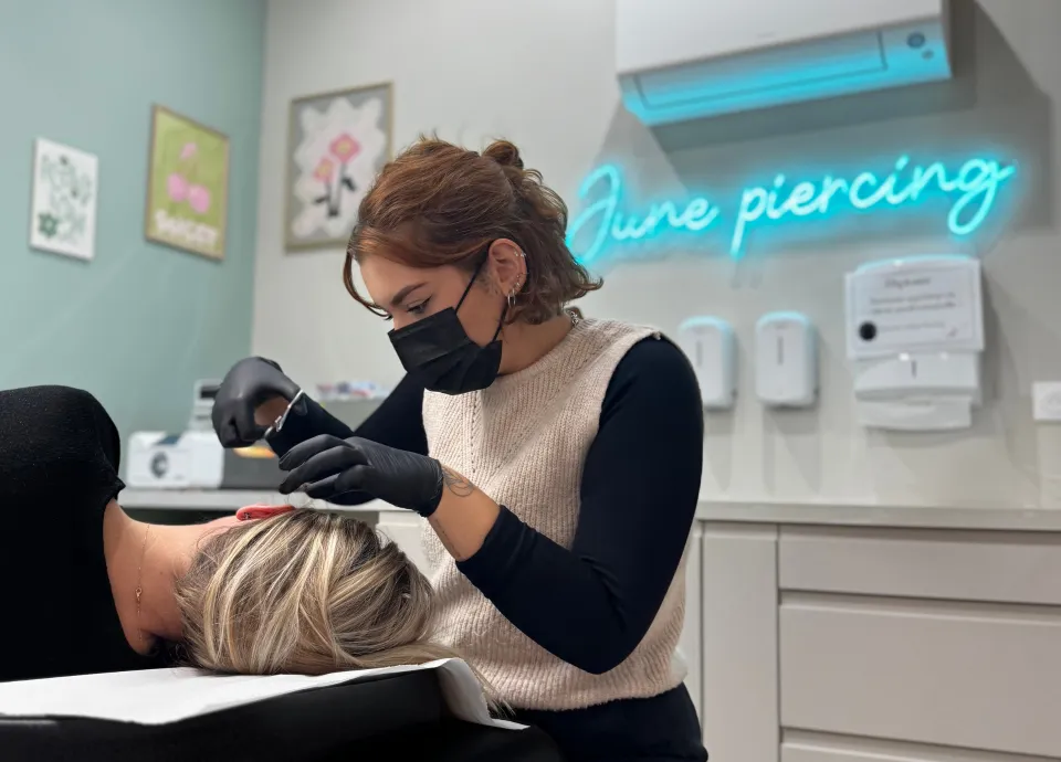 Une pierceuse portant un masque et des gants noirs réalise un piercing sur l'oreille d'une cliente allongée, devant un néon "June Piercing".