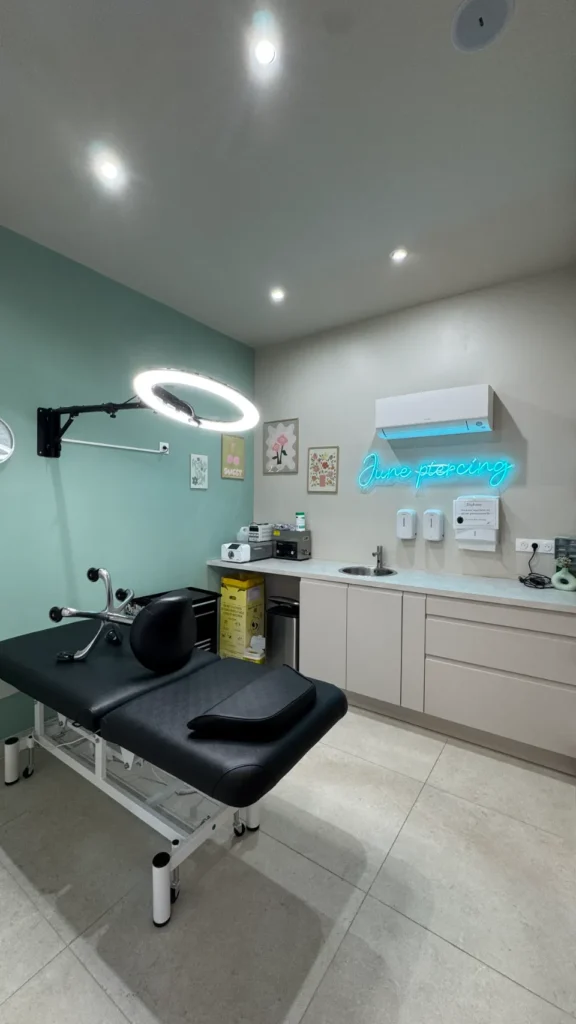 Salle de piercing moderne avec un lit de procédure noir, un lavabo, une enseigne au néon 'June Piercing' et un éclairage professionnel.