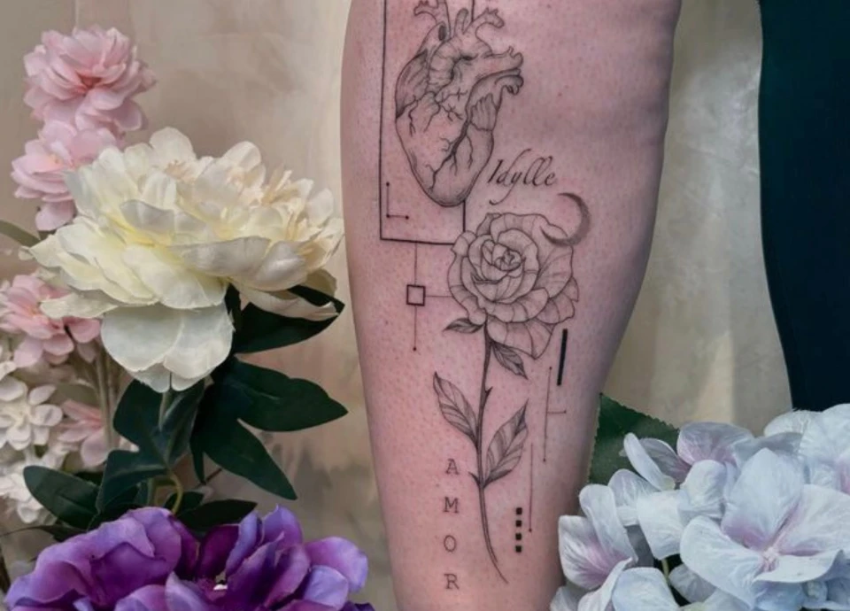 Tatouage sur la jambe représentant un cœur anatomique stylisé, une rose dessinée, la lune, et le mot "amour" écrit à la verticale. Le motif est encadré par des lignes géométriques.