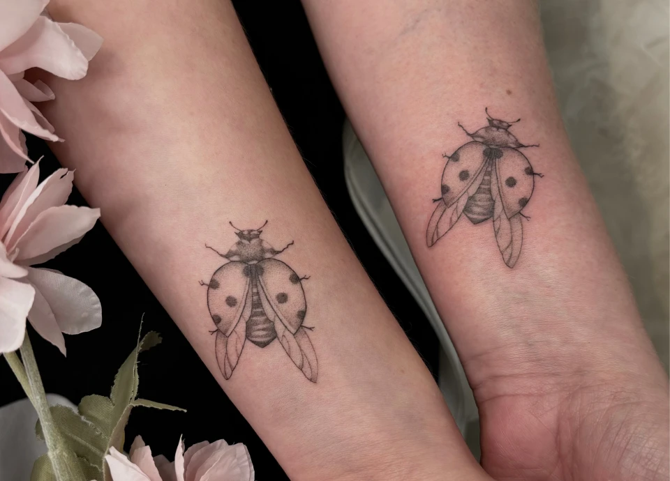 Deux tatouages de coccinelles réalistes, dessinés à l'encre noire, sont visibles sur les avant-bras d'une personne.