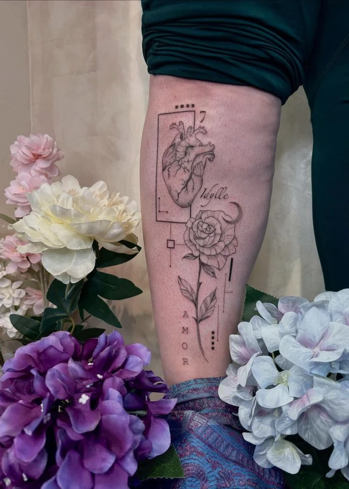 Tatouage sur une jambe représentant un cœur anatomique, une rose, les mots "Idylle" et "Amor" et des formes géométriques.