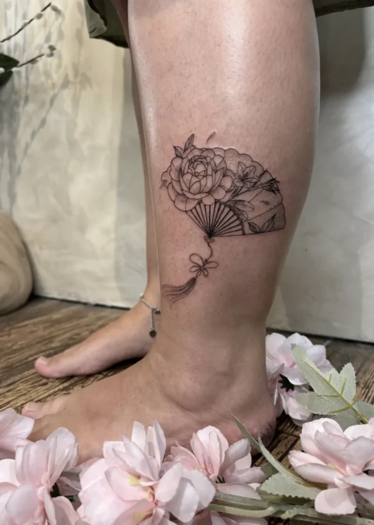 Tatouage en noir et blanc d'un éventail orné de pivoines et de petites fleurs, avec un gland, situé sur la cheville d'une femme.