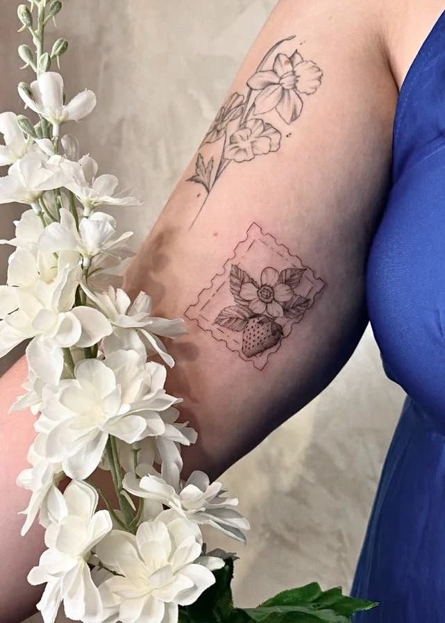 Tatouage d'une fraise et d'une fleur dans un cadre rappelant un timbre-poste, sur un bras orné de fleurs blanches.
