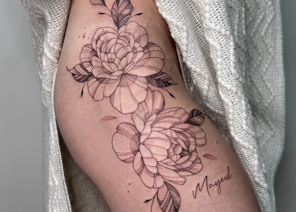 Tatouage de deux fleurs stylisées aux pétales délicats, sur la cuisse d'une personne, avec un nom manuscrit en bas à droite.