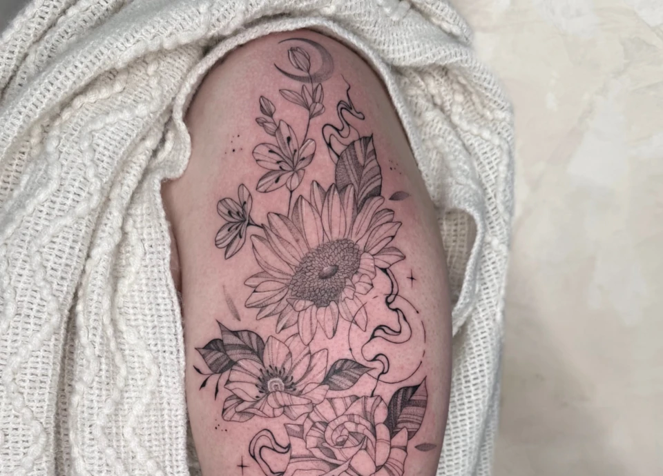 Tatouage détaillé d'un tournesol, de petites fleurs, d'une rose et d'un croissant de lune, orné de volutes et d'étoiles.