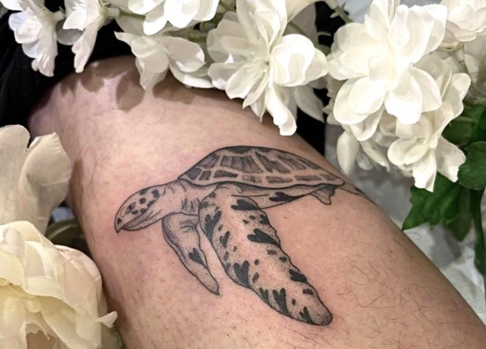 Tatouage réaliste en noir et blanc d'une tortue sur un bras, entouré de fleurs blanches.