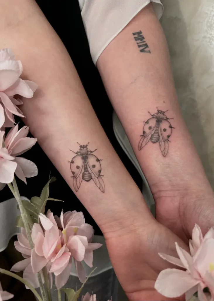 Deux tatouages réalistes de coccinelles aux ailes déployées, sur les avant-bras de deux personnes, entourés de fleurs roses.