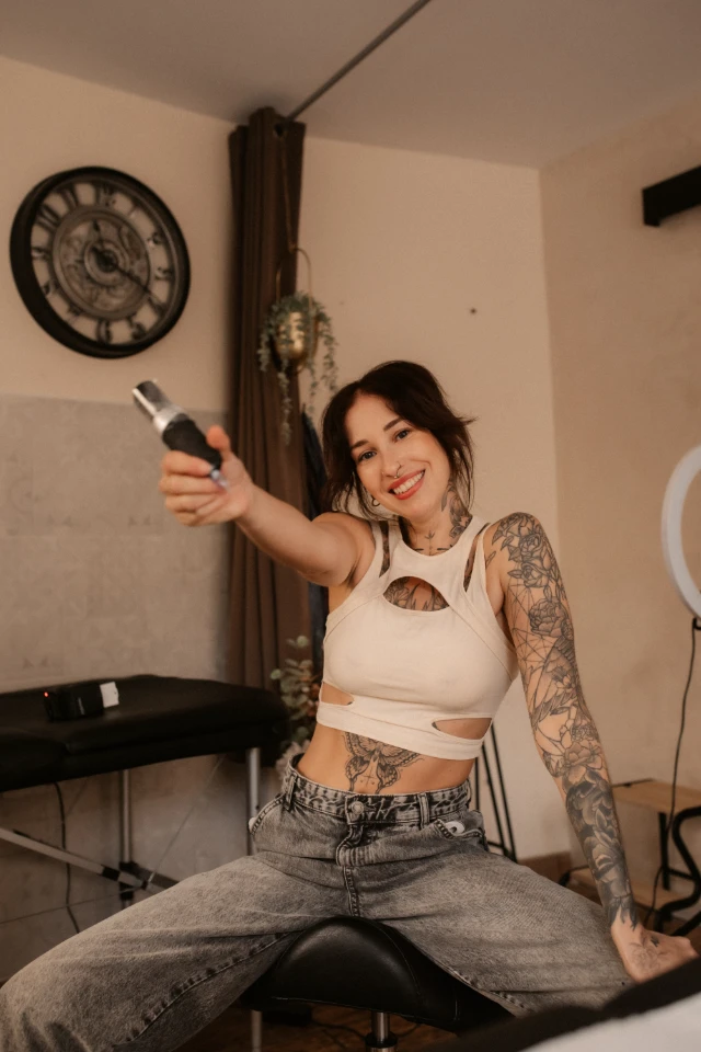 Tatoueuse aux bras couverts de dessins tenant un dermographe, souriant à la caméra, assise devant une table de massage.