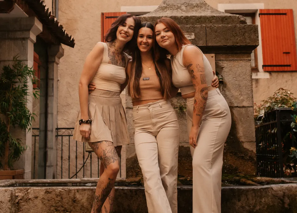 Trois jeunes femmes souriantes et tatouées, habillées de tenues beiges, posant ensemble devant une fontaine en pierre et un bâtiment.